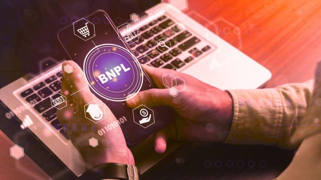 illustration of a man holding a phone with a BNPL option in his shopping cart on his smart phone as he sits at his desk with a laptop on it.