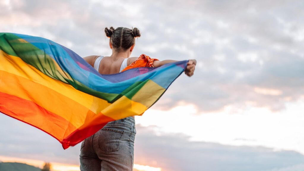 A photo of the back of a person who is holding a pride flag and looking at the sunset gettyimages-1327194458