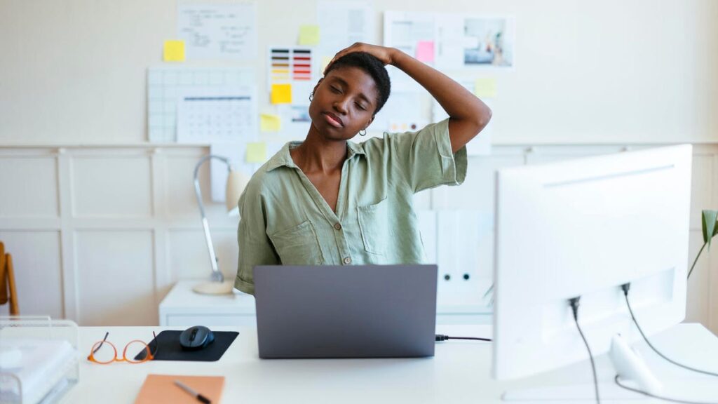 A person with short black hair and a light green button-up blouse stretching their neck at a white desk in front of a silver laptop.