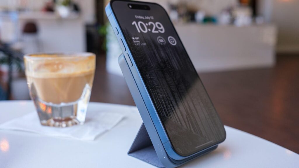My Favorite Portable iPhone Charger Is 35% Off in All Colors iPhone connected to a slim portable charger on a stand. They're both on a table in a brightly lit cafe along with a cortado in a glass.