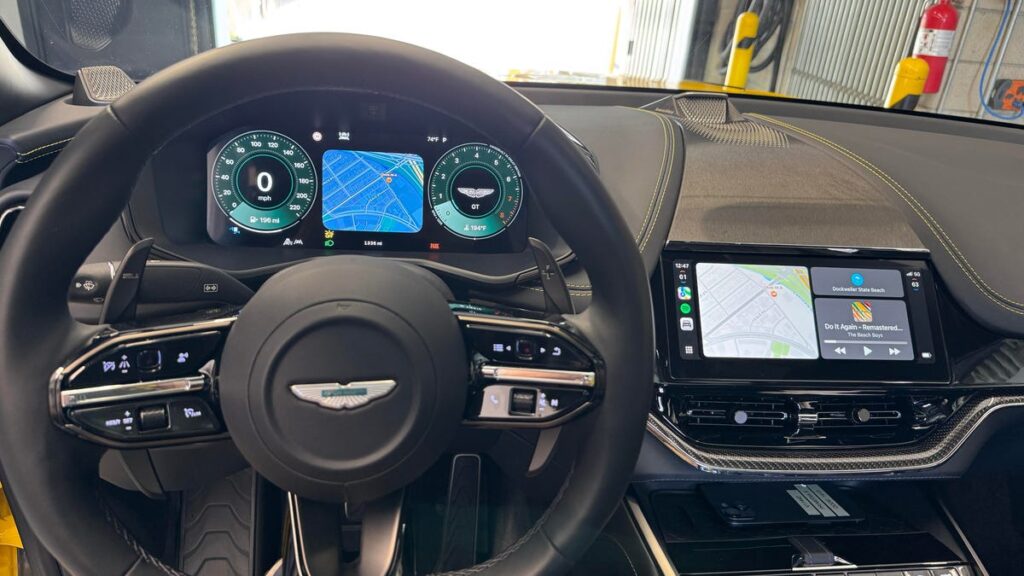 A view of the wheel and dashboard of an Aston Martin crossover SUV, showing different maps and controls on the screens in the center and behind the wheel.
