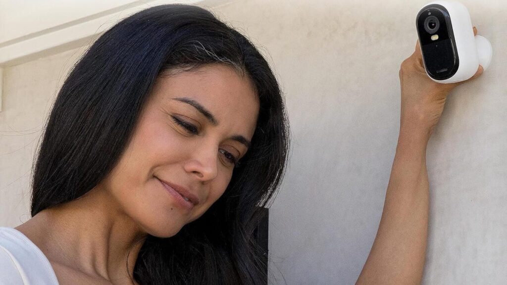 A woman smiles at a phone as she reaches up to an Arlo Essential cam on a wall.