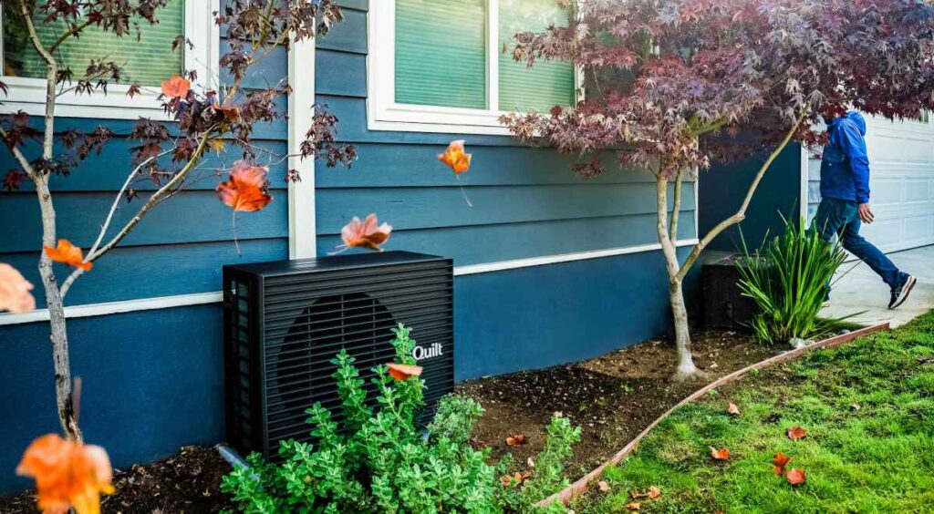 How Quilt solved the heat pump’s biggest challenge A dark grey heat pump sits outside a blue home.