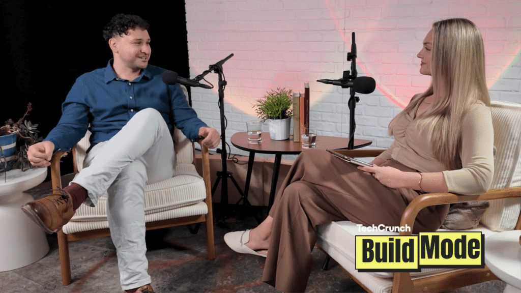Two people seated in armchairs during a podcast interview, speaking into microphones across a small table with water glasses and a plant, with a “TechCrunch Build Mode” logo overlay in the corner.