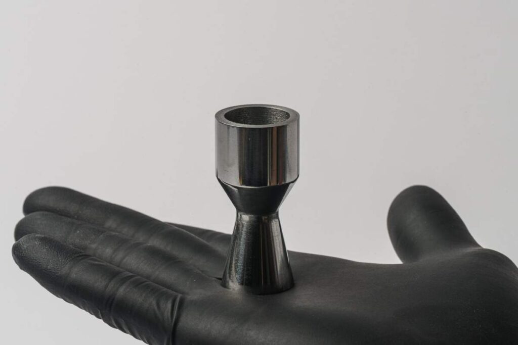 A gloved hand holds a metal part shaped like a rocket nozzle.